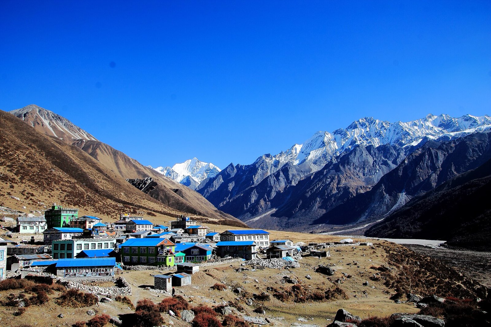 A cluster of blue-roofed houses on a valley floor beneath snow-covered peaks in Rasuwa
